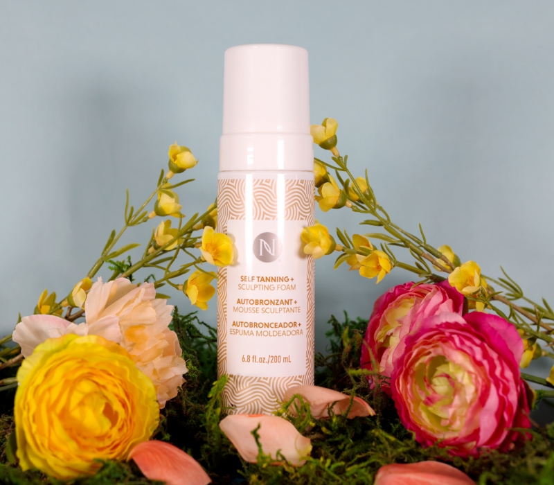 Neora's 3-in-1 Self Tanning + Sculpting Foam sitting in a bed of spring flowers.
